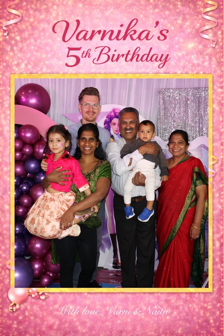 Family photo from Varnikas 5th birthday celebration with six people posing against pink balloons and sparkly backdrop
